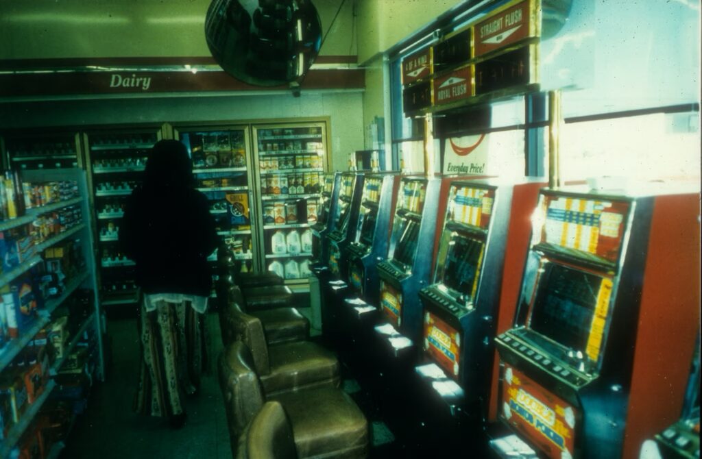 A digitization of a photo slide showing a 1970s convenience store with a row of slot machines lining the outer wall of the store with windows out to the pumps above the machines. A person is walking away from the camera to the back built-in refrigeration units.
