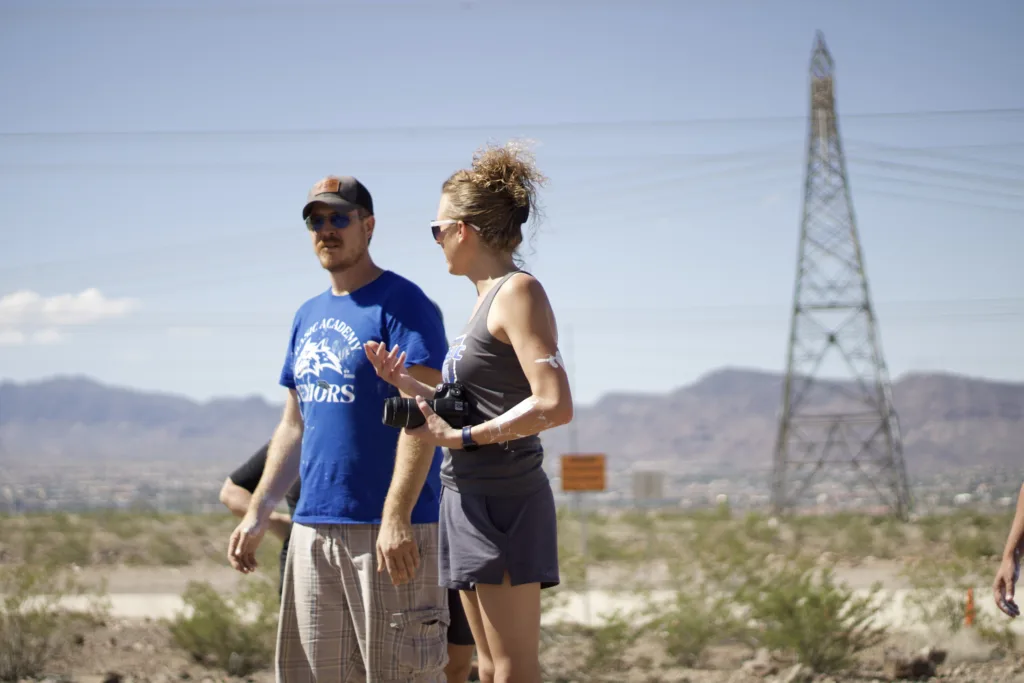 Two teachers, a man and woman, having a conversation. They are standing outside in the desert and she is holding a camera.