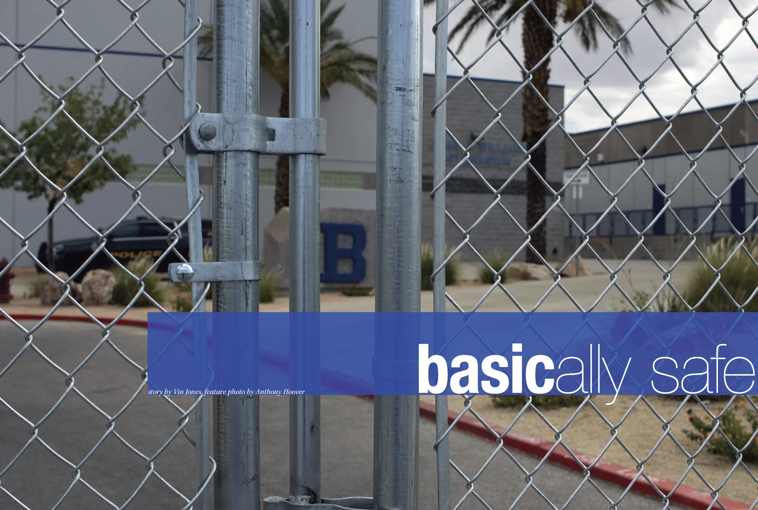 A photo of a chain-link fence with a blue and white sign that says "basically" in lowercase letters. In the background, a school building is partially visible.