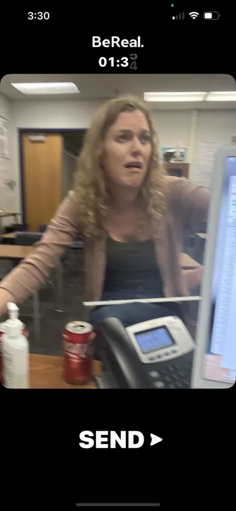 A blonde woman in a tan cardigan and black top sitting at a desk in a classroom, captured in a BeReal post with a "01:34" timer overlay.