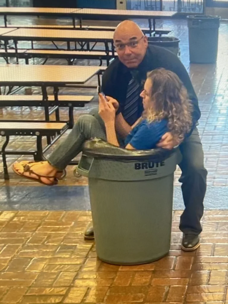 A woman sits inside a large gray trash can in a school cafeteria while looking at her smartphone, as a man in a black shirt and tie poses playfully behind her.