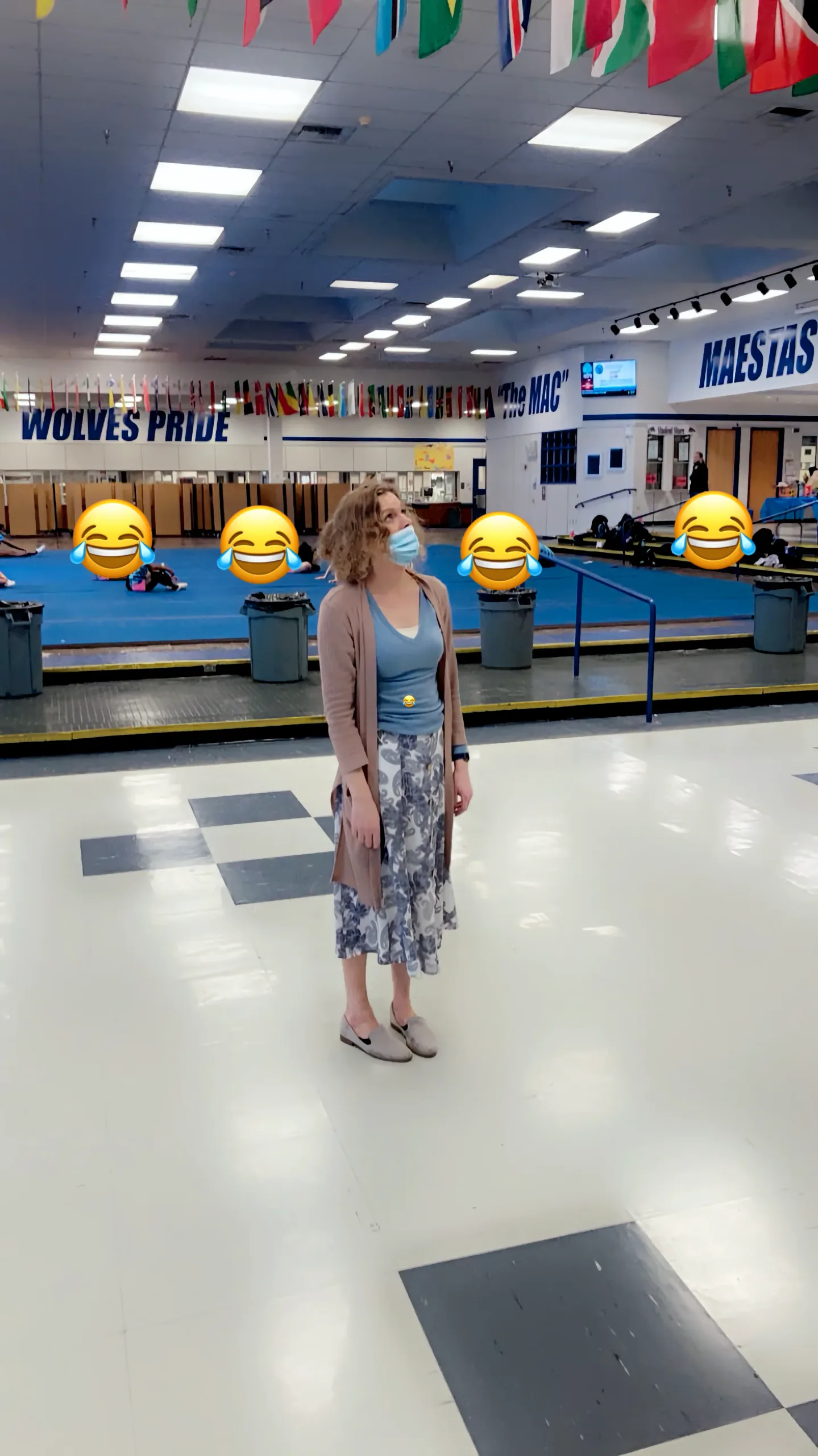 I am standing in the center of a large, empty high school cafeteria/gym during the pandemic. I’m wearing a face mask, a long cardigan, and a patterned skirt. In the background, there are "Wolves Pride" and "The MAC" signs, blue athletic mats, and several large "laughing crying" emojis covering the faces of people in the distance.